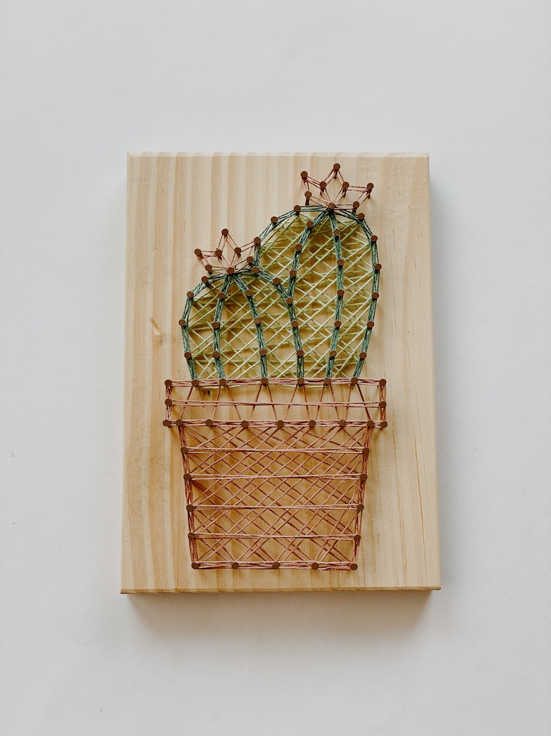 May include: A string art cactus with green and brown yarn, in a terracotta pot, on a wooden background.