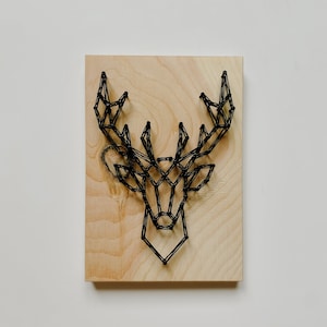 May include: A black string art deer head mounted on a wooden plaque. The deer head is made of black string wrapped around nails hammered into the wood.