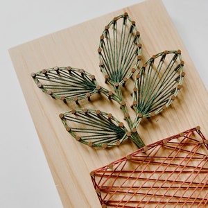 Puede incluir: Arte de hilo hecho a mano con un diseño de planta. La obra de arte está hecha sobre una tabla de madera clara. La planta tiene hojas verdes y una maceta marrón rojizo, todo creado con hilo y pequeños clavos. Una pieza de decoración del hogar única.