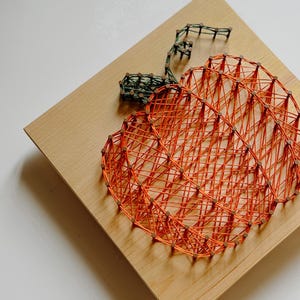 K&ouml;nnte beinhalten: Orangefarbener String-Art-K&uuml;rbis auf einem Holzgrund. Der K&uuml;rbis besteht aus orangefarbenem Garn, das um N&auml;gel gewickelt ist, die in das Holz geschlagen wurden. Der Stiel ist aus gr&uuml;nem Garn.
