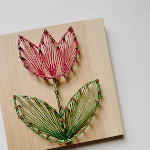 Peut inclure: Une œuvre d'art en fil représentant une tulipe. Les pétales sont en fil rouge, la tige est verte et les feuilles sont également vertes. L'œuvre est montée sur une planche de bois carrée, créant une esthétique rustique. Dimensions: environ 20 cm x 20 cm.
