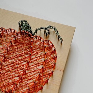 K&ouml;nnte beinhalten: Orangefarbener String-Art-K&uuml;rbis mit gr&uuml;nem Stiel auf einem Holzgrund.
