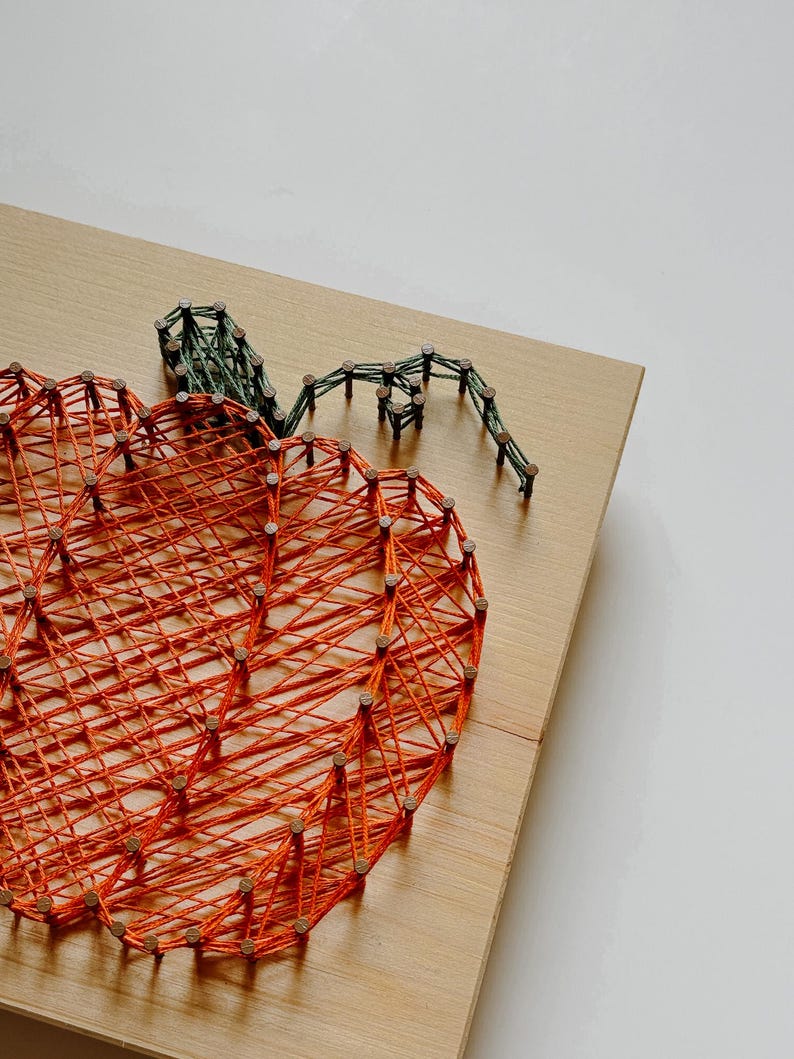 K&ouml;nnte beinhalten: Orangefarbener String-Art-K&uuml;rbis mit gr&uuml;nem Stiel auf einem Holzgrund.