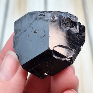 May include: A faceted, black tourmaline crystal. The gemstone has a glossy, reflective surface with sharp edges and a complex geometric shape. The crystal is held in a hand against a light-colored wooden background.