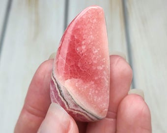 May include: A polished rhodochrosite gemstone with a teardrop shape, featuring vibrant shades of pink and coral with distinct white and dark grey banded layers. The surface displays a crystalline texture with a smooth, glossy finish throughout.