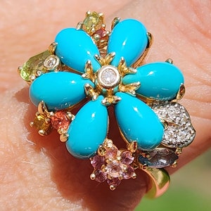 May include: A gold ring featuring a flower design with six smooth, teardrop-shaped turquoise petals surrounding a central round diamond. The setting includes additional small accent stones in shades of pink, yellow, and green, along with a leaf-shaped element encrusted with tiny white diamonds.
