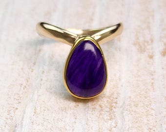 May include: A gold ring with a teardrop-shaped, deep purple gemstone. The ring's band curves upwards on either side of the gemstone, creating a unique design. The gemstone is set in a gold bezel. A stylish piece of jewellery.