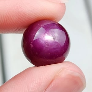 May include: A round, polished, deep magenta gemstone. The gem is held between two fingers, showcasing its smooth, reflective surface. The stone's color is a rich, saturated hue, with a subtle play of light.
