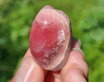 May include: A polished rhodochrosite gemstone with a smooth, rounded shape. The stone displays vibrant shades of deep pink and soft rose, featuring distinct concentric banding patterns in lighter white and cream tones across its surface.