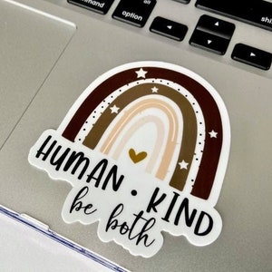 May include: A sticker with a rainbow design in shades of brown and beige with a small gold heart in the center. The text "HUMAN. KIND be both" is written below the rainbow.
