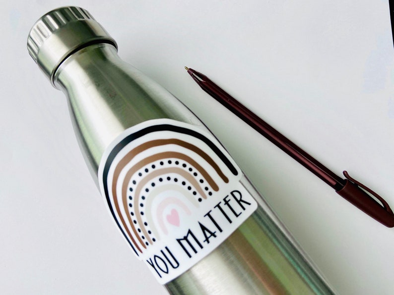 May include: A stainless steel water bottle with a sticker featuring a rainbow with brown, beige, and black stripes and a pink heart. The sticker also includes the text "You Matter".