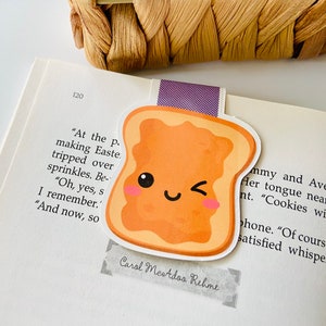 May include: A cute cartoon bookmark shaped like a slice of toast with peanut butter. The toast has a smiling face with a wink and rosy cheeks. The bookmark is on a page of a book with text that reads "At the p making Easte tripped over sprinkles. Be- "Oh, yes, s I remember. "And now, so Carol Mestdoo Rehme mmy and Avery Her tongue nearly nt. "Cookies with phone. "Of course, satisfied whisper."