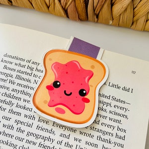 May include: A cute cartoon illustration of a slice of toast with a pink jelly spread. The toast has a happy face with closed eyes and a smile. The toast is a bookmark.