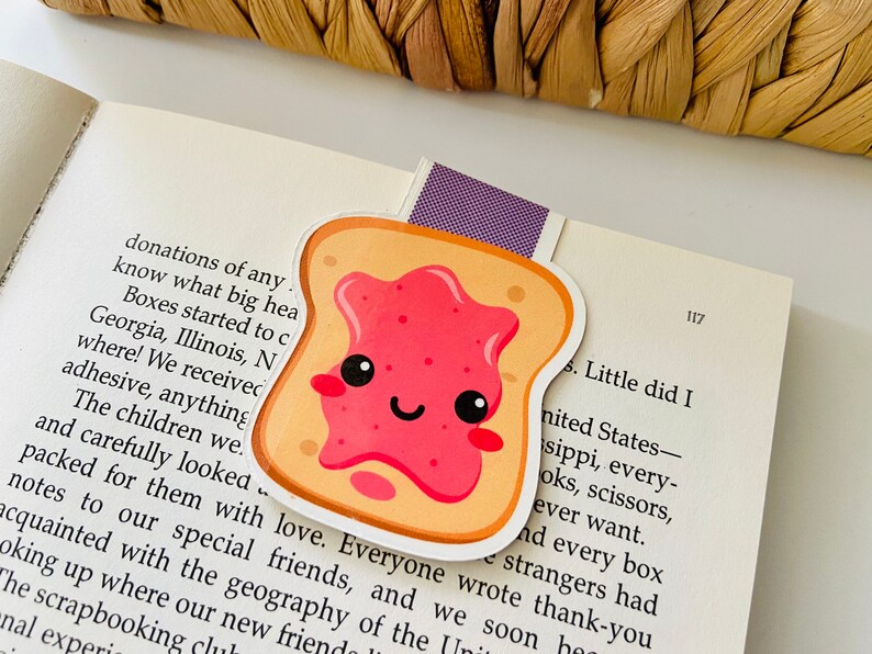 May include: A cute cartoon illustration of a slice of toast with a pink jelly spread. The toast has a happy face with closed eyes and a smile. The toast is a bookmark.