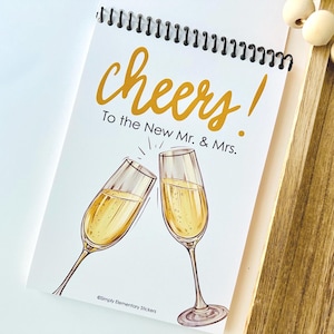 May include: A spiral-bound notepad with the word "cheers!" in gold script above the phrase "To the New Mr. & Mrs." Two champagne flutes are illustrated below. The notepad is white with a black spiral binding.