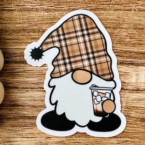 May include: A brown and white plaid gnome holding a brown and white cup with white marshmallows. The gnome has a white beard and black shoes.