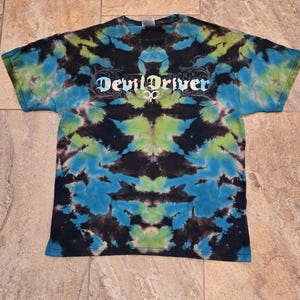 Puede incluir: Camiseta teñida con un patrón de tie-dye en azul, verde y negro. La camiseta tiene el texto "DevilDriver" en una fuente estilizada en el pecho. La camiseta está colocada sobre una superficie embaldosada.