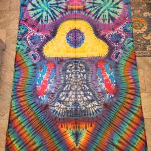 May include: A vibrant tie-dye textile featuring a kaleidoscope of colors. The design includes concentric patterns in shades of blue, purple, yellow, orange, and green. The central motif is a symmetrical, cloud-like shape with intricate details.