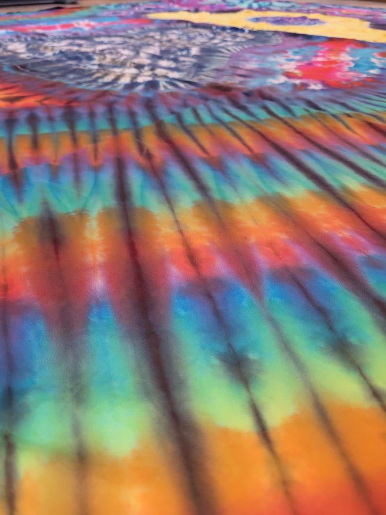 May include: A vibrant tie-dye textile featuring a spectrum of colors, including orange, red, blue, and green. The design has a striped pattern with dark brown lines. The fabric appears to be spread out, with a section of blue and white tie-dye in the background.