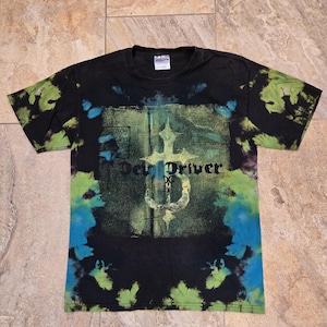 Puede incluir: Camiseta negra teñida con un diseño gráfico. La camiseta presenta un diseño central con el texto "Dev Driver" en fuente gótica y un símbolo. La camiseta tiene acentos teñidos de verde y azul.