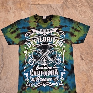 Puede incluir: Camiseta teñida con tinte anudado en tonos verdes, azules y negros. La camiseta presenta un gráfico blanco con las palabras "DEVILDRIVER" sobre herramientas cruzadas y "Genuine CALIFORNIA Groove" debajo.