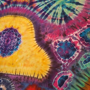 May include: A vibrant, colorful tie-dye textile with a psychedelic design. The pattern features concentric circles and spiral shapes in shades of yellow, purple, blue, pink, and green. The fabric has a soft, flowing appearance, suggesting it could be a blanket or scarf.