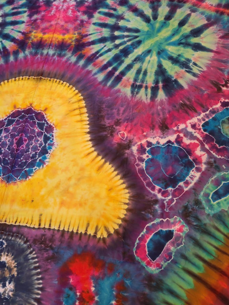 May include: A vibrant, colorful tie-dye textile with a psychedelic design. The pattern features concentric circles and spiral shapes in shades of yellow, purple, blue, pink, and green. The fabric has a soft, flowing appearance, suggesting it could be a blanket or scarf.
