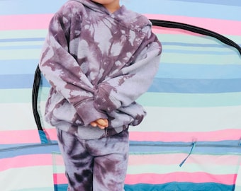 May include: A young child wearing a purple tie-dye hoodie and matching pants. The child is standing in front of a colorful striped background.