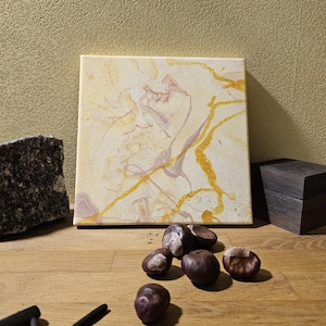 Op de afbeelding: Een vierkant canvas schilderij met abstracte wervelingen van crème, lichtroze en metallic goudkleurige verf. Links staat een ruwe, donkergrijze granieten steen, terwijl er voor het schilderij een klein donkerhouten doosje en enkele bruine kastanjes liggen.