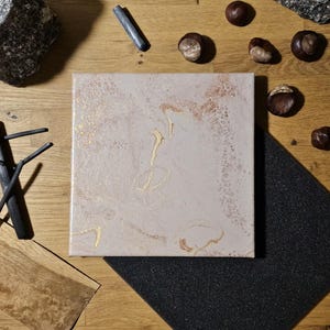 Op de afbeelding: Een vierkant canvas met een lichtbeige basis en wervelende patronen van metallic goud- en koperkleurige verf. Het oppervlak heeft een getextureerd, gemarmerd uiterlijk met subtiele variaties in kleurintensiteit en vloeiende, organische vormen.