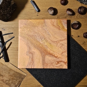 Op de afbeelding: Een vierkant canvas met een abstracte acryl gietschildering met wervelende tinten perzik, crème en metallic goud. Het oppervlak vertoont een gemarmerde textuur met variërende verfdichtheden, wat een vloeiend en organisch patroon creëert.
