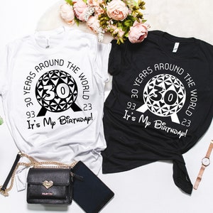 May include: Two t-shirts, one white and one black, with a graphic design of a black and white Spaceship Earth icon from Epcot. The text on the shirts reads "30 Years Around The World 93. 23 It's My Birthday!"