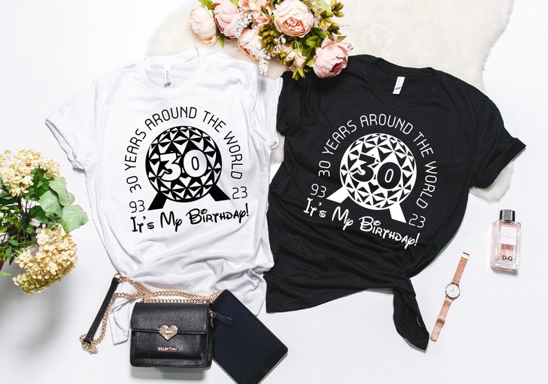 May include: Two t-shirts, one white and one black, with a graphic design of a black and white Spaceship Earth icon from Epcot. The text on the shirts reads "30 Years Around The World 93. 23 It's My Birthday!"