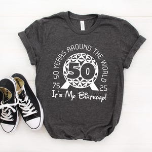 May include: A dark grey t-shirt with a white graphic of a sphere with the text "50 Years Around The World 75 50 25 It's My Birthday!" printed on it.