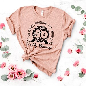 May include: A pink t-shirt with a black graphic design of a sphere with the number 50 inside. The text around the sphere reads "50 Years Around The World 75 25 It's My Birthday!"