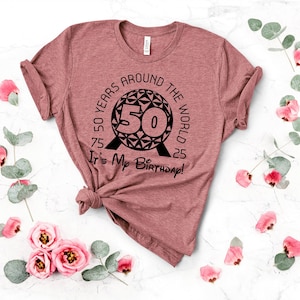 May include: A pink t-shirt with a black graphic of a sphere with the text "50 Years Around The World" and "It's My Birthday!" printed on it.