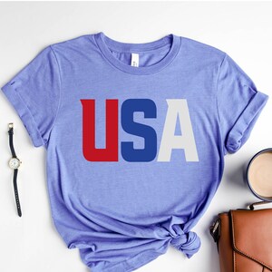 Peut inclure: Un t-shirt bleu clair avec le mot "USA" imprimé en rouge, blanc et bleu. Le t-shirt est plié et placé sur une surface blanche, à côté d'un plateau noir, de lunettes, d'une montre, d'un sac à main et d'une tasse de café.