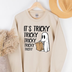 May include: Beige sweatshirt with the text "IT'S TRICKY TRICKY TRICKY TRICKY TRICKY" in black, along with a white ghost holding a brown paper bag. The sweatshirt is hanging on a wooden hanger, with a straw hat in the background. Halloween themed apparel.