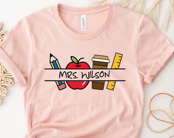May include: A light pink t-shirt featuring a teacher-themed graphic. The design includes a pencil, a red apple, a coffee cup, and a ruler, with the name "MRS. WILSON" in the center. The shirt is made of soft material.
