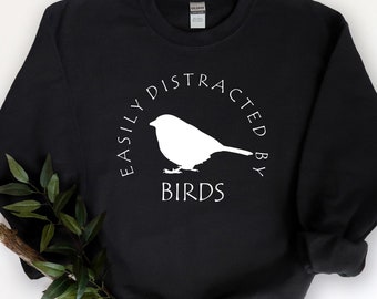 May include: Black sweatshirt with the text "EASILY DISTRACTED BY BIRDS" in a white circular design. A white bird silhouette is in the center of the circle. The sweatshirt is hanging on a wooden hanger.