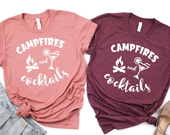 May include: Two t-shirts with the text "Campfires and Cocktails" in white lettering. The shirts are in a pink and burgundy color. The text is accompanied by a campfire and a cocktail glass graphic.
