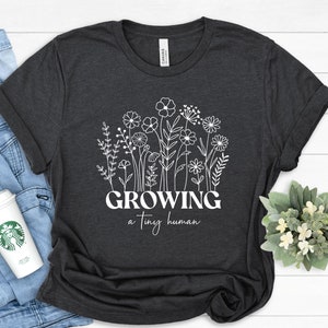 May include: A dark gray t-shirt with a white graphic of flowers and the text "GROWING a tiny human".