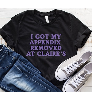 May include: Black t-shirt with purple text that reads "I got my appendix removed at Claire's". The shirt is laid out on a white wooden surface with a pair of gray canvas shoes and blue jeans.