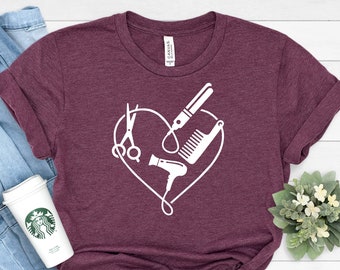 May include: A maroon t-shirt with a white graphic of hairdressing tools arranged in a heart shape. The tools include scissors, a hair dryer, a comb, and a curling iron.