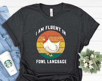 May include: A dark grey t-shirt with a vintage-style graphic of a chicken in front of a sunset. The text "I am fluent in fowl language" is printed below the graphic.
