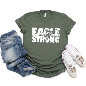 Op de afbeelding: Een groen T-shirt met witte tekst die "Eagle Strong" zegt. De tekst is gelaagd over een adelaargrafiek. Het shirt ligt plat op een wit oppervlak met een blauwe spijkerbroek en witte sneakers.