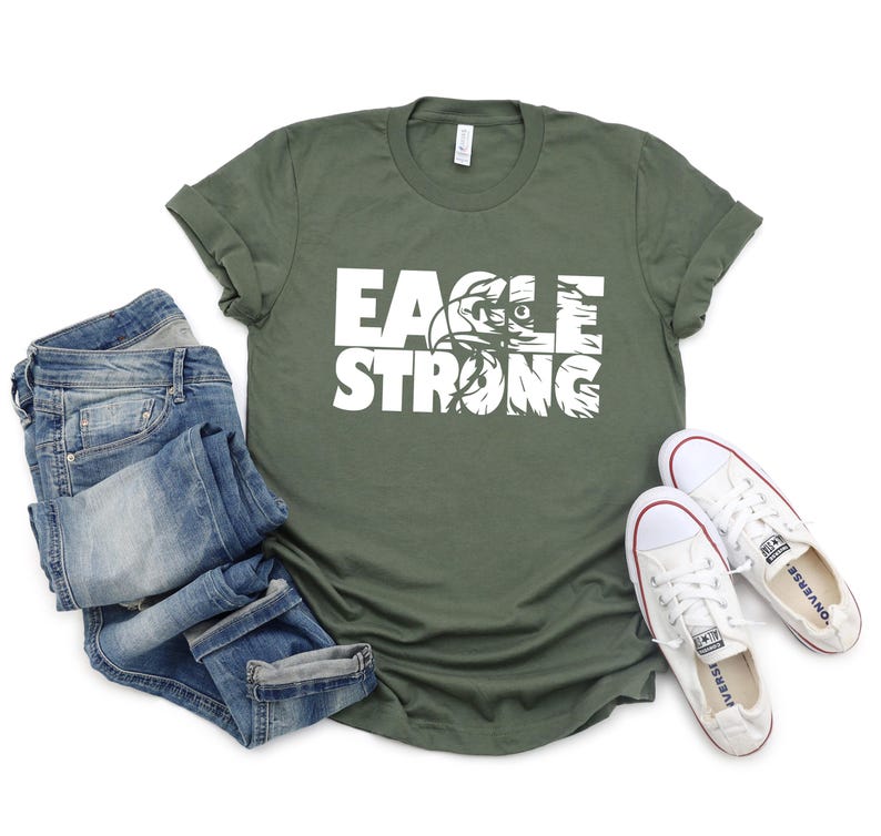 Op de afbeelding: Een groen T-shirt met witte tekst die "Eagle Strong" zegt. De tekst is gelaagd over een adelaargrafiek. Het shirt ligt plat op een wit oppervlak met een blauwe spijkerbroek en witte sneakers.