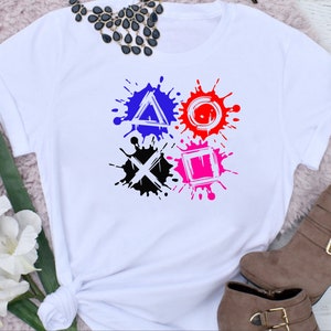 May include: White t-shirt with a graphic of the PlayStation controller symbols in black, blue, red, and pink paint splatters.
