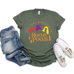 Puede incluir: Una camiseta verde con el texto "It's just a bunch of Hocus Pocus" en naranja y amarillo. Tres brujas est&aacute;n representadas en el dise&ntilde;o, con su cabello en morado, naranja y amarillo.
