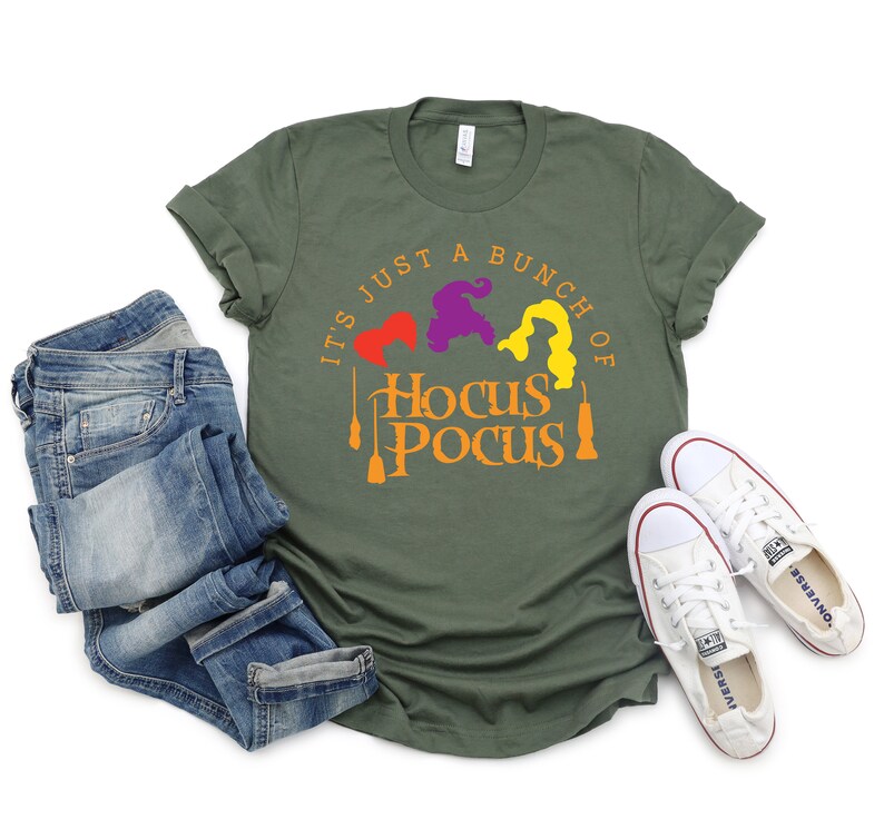Puede incluir: Una camiseta verde con el texto "It's just a bunch of Hocus Pocus" en naranja y amarillo. Tres brujas est&aacute;n representadas en el dise&ntilde;o, con su cabello en morado, naranja y amarillo.